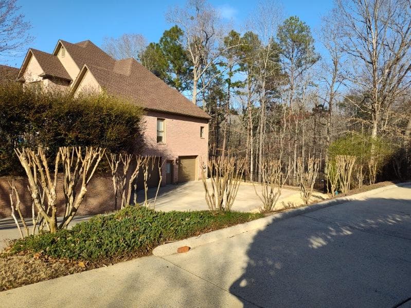 Two-story brown house surrounded by trees and trimmed shrubs on a sunny day.