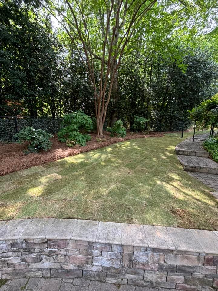 Lush green garden area with newly laid sod, surrounded by trees and a stone wall.