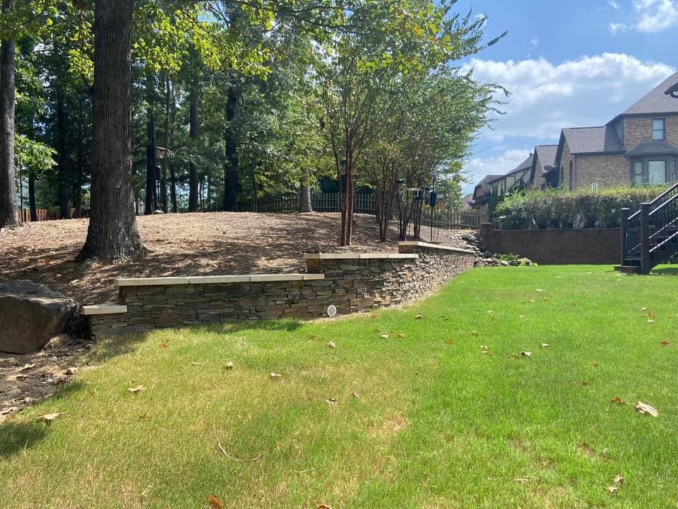 Landscape featuring a stone wall, green lawn, trees, and homes in the background. Ideal for outdoor living.