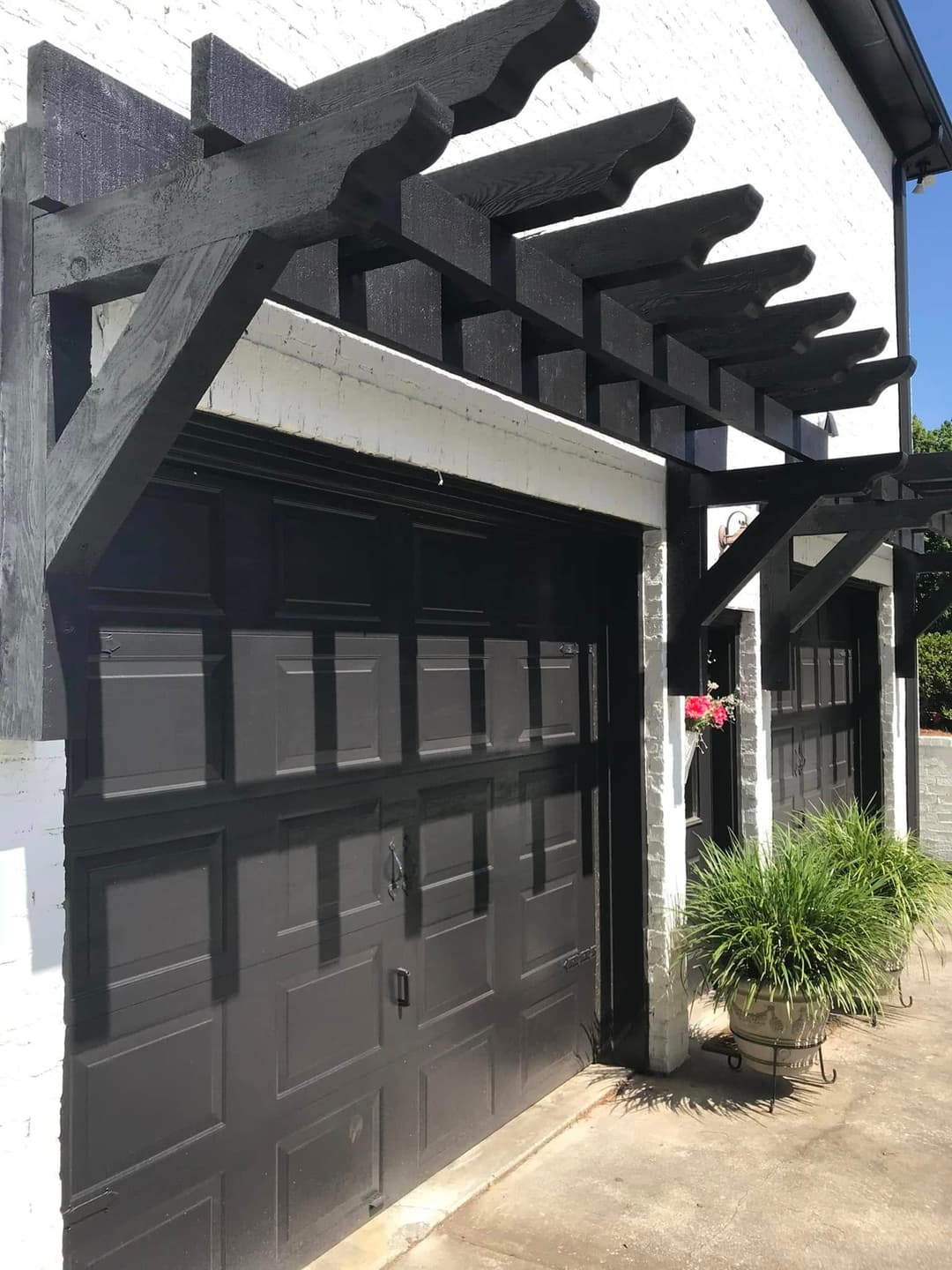 Black garage doors under a wooden pergola with green plants and flowers by a white house.