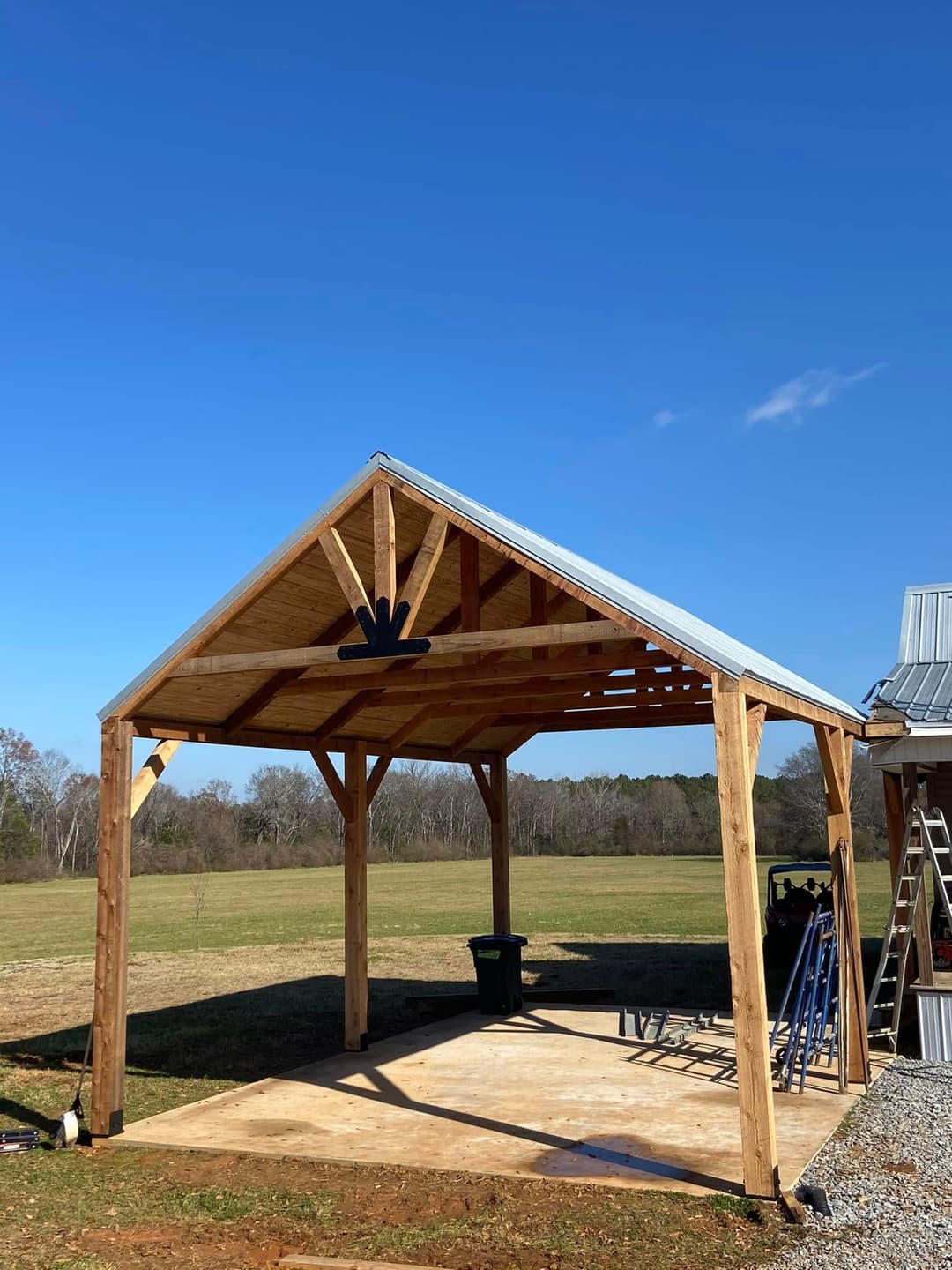 Open wooden pavilion with a metal roof in a field under a clear blue sky.