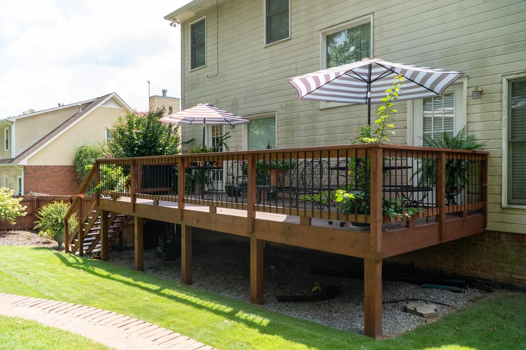 Wooden deck with planters and umbrellas, surrounded by grass and a brick path.