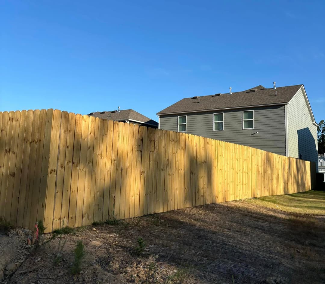 New wooden fence enclosing backyard with two houses in the background and clear blue sky.