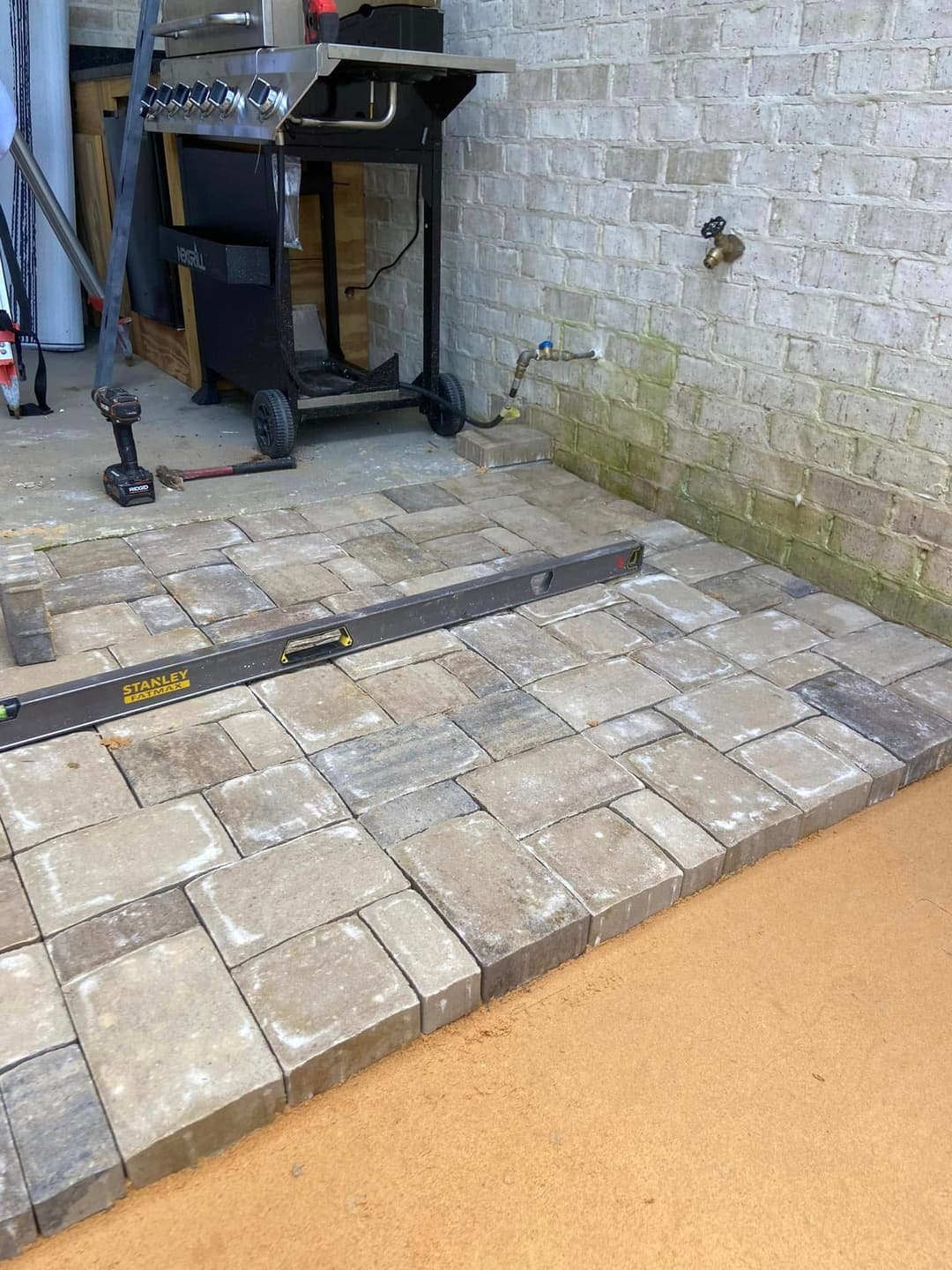 Stone patio installation in progress with leveling tool, outdoor grill, and unfinished area.