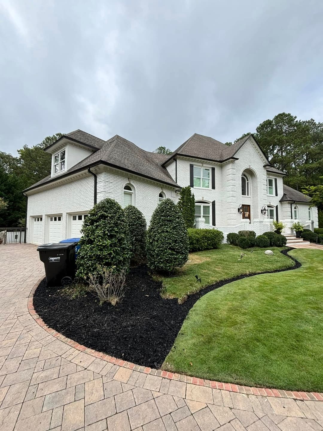 Elegant white brick house with landscaped yard, circular driveway, and black trash can.