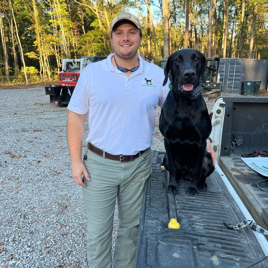 Smiling man in a white shirt stands next to a black Labrador Retriever on a truck bed.