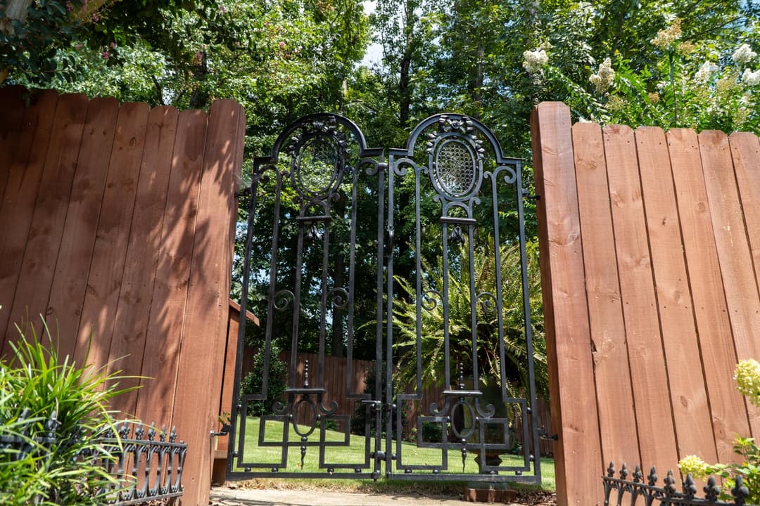 Wrought iron garden gate opening to a lush green backyard surrounded by wooden fencing.