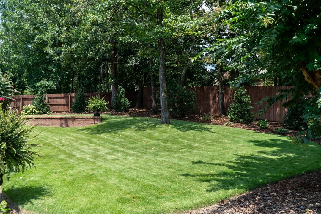 Lush green backyard with manicured lawn, trees, and wooden privacy fence.