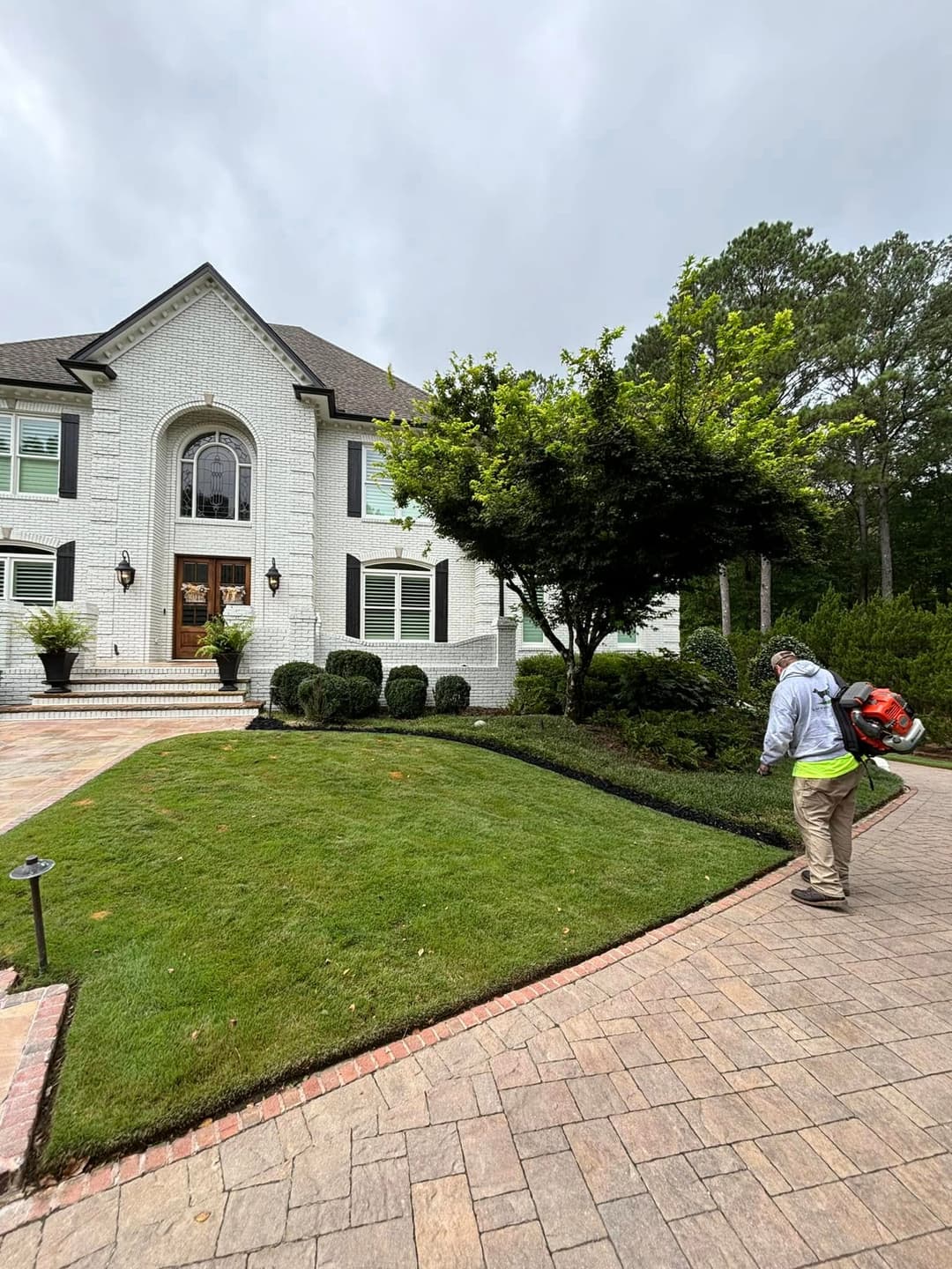 Landscape maintenance with a man using a blower near a well-kept white brick house.