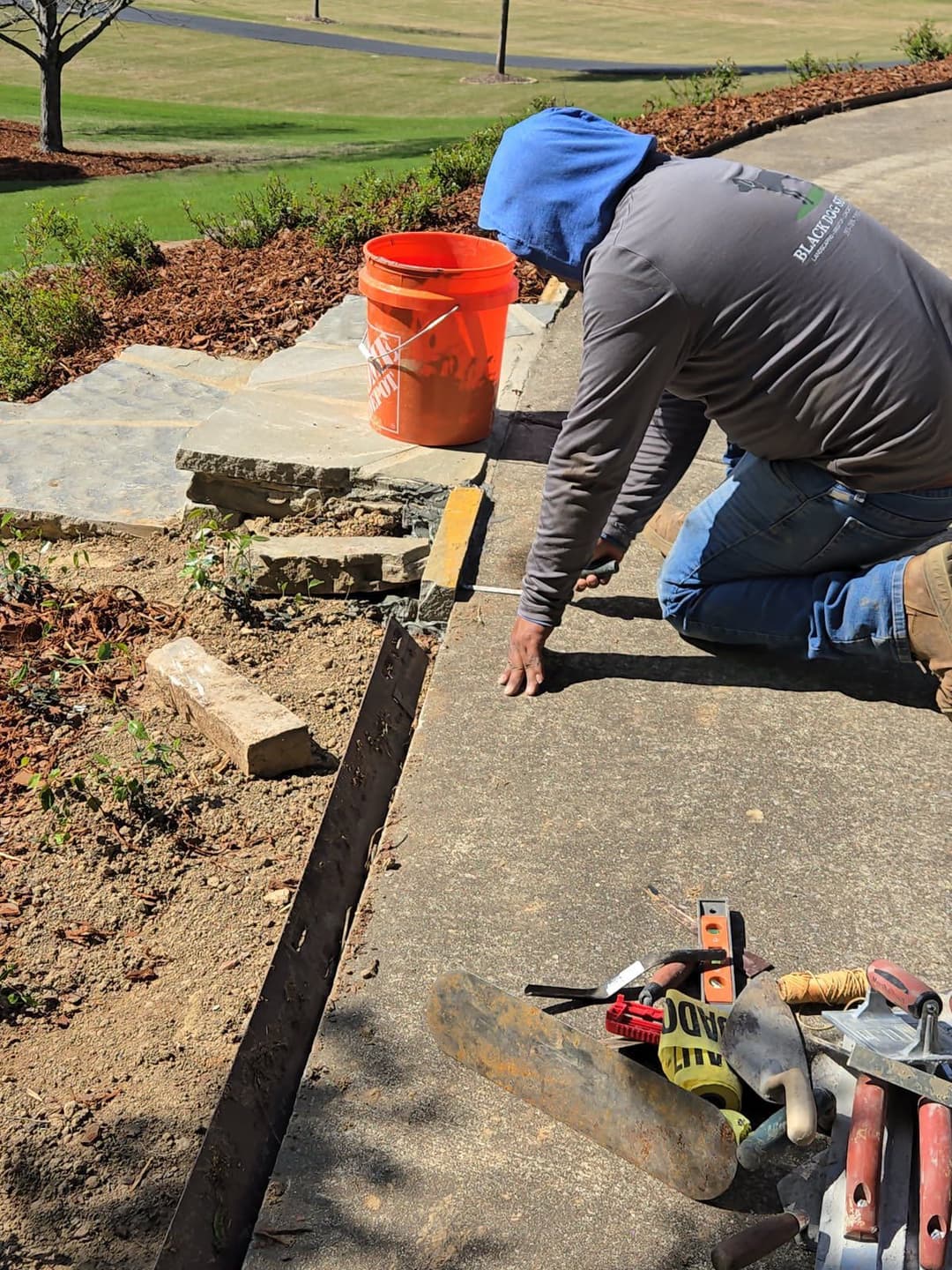 Person working on landscape edging with tools and a bucket on a sunny day.