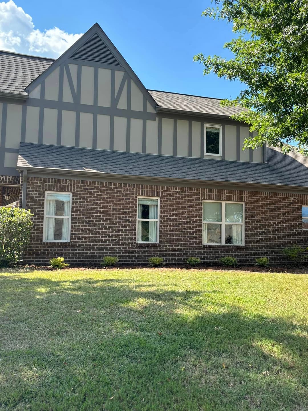 Brick house with gray siding, steep roof, and well-maintained lawn under a blue sky.