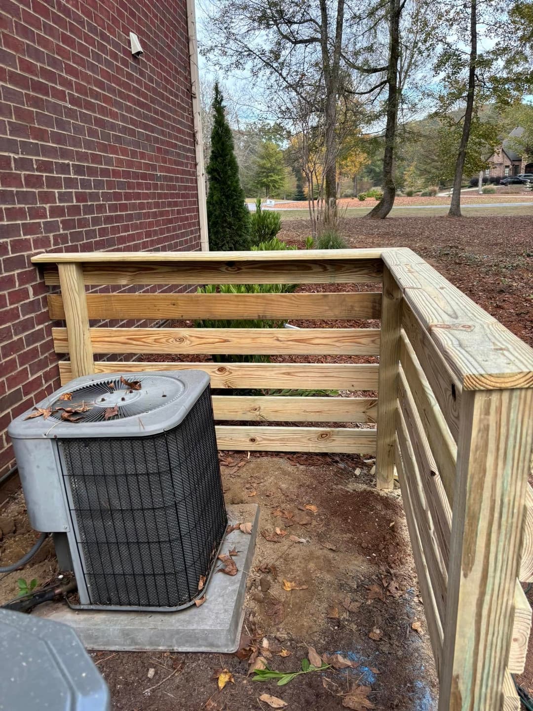Wooden fence enclosure around an air conditioning unit beside a brick building.