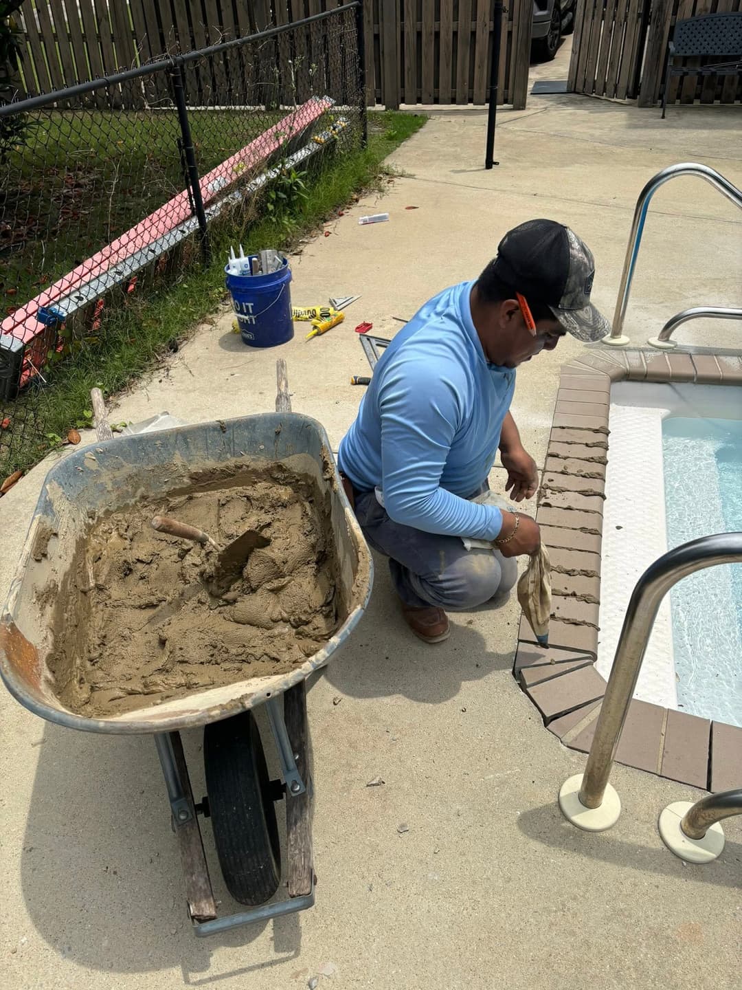Worker repairing poolside tiles with a wheelbarrow of mortar in a backyard setting.