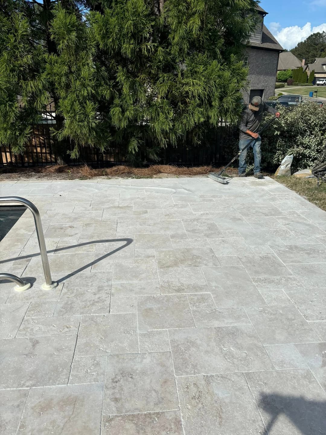 Worker finishing a stone patio around a pool, surrounded by greenery and a privacy fence.