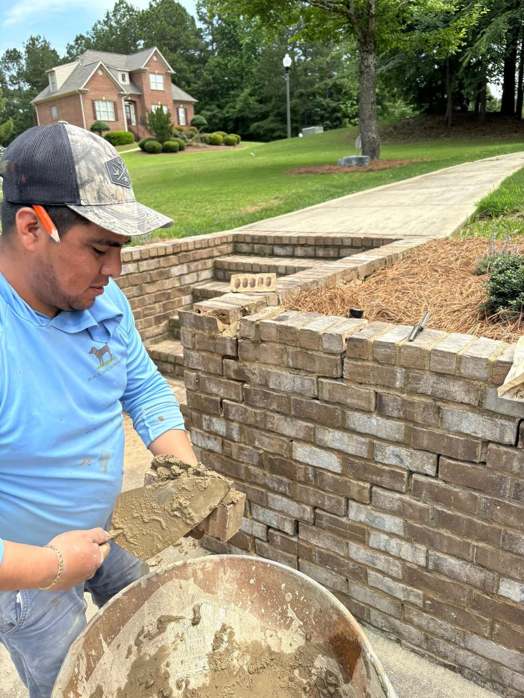 Mason applying mortar while building a stone wall in a landscaped yard.