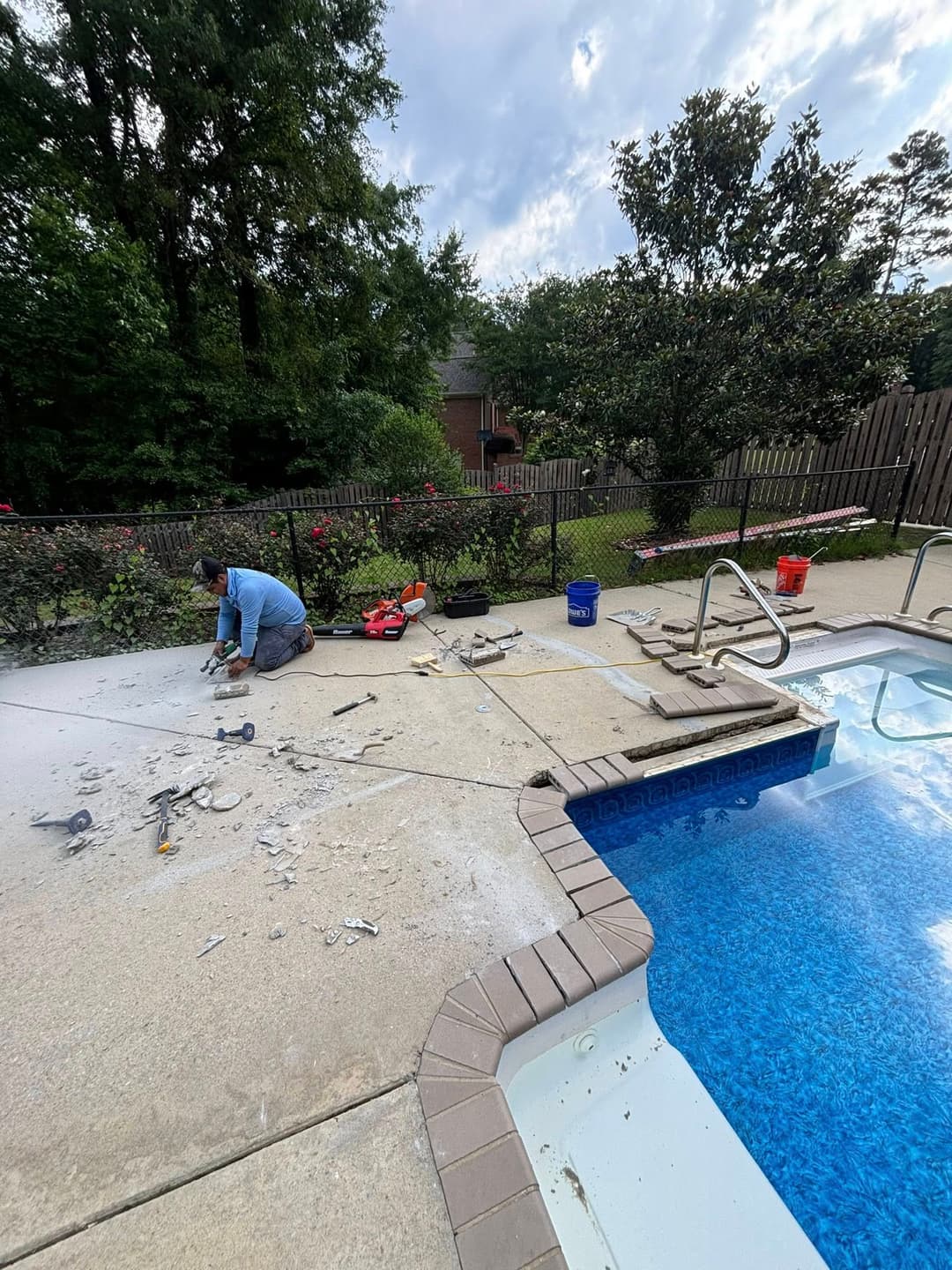 Worker renovating pool deck with tools beside a refreshing blue swimming pool and lush landscaping.