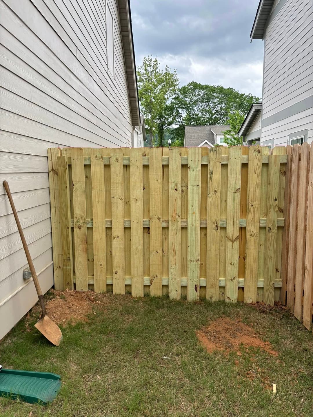 New wooden fence installation between houses with a shovel on the ground and grass.
