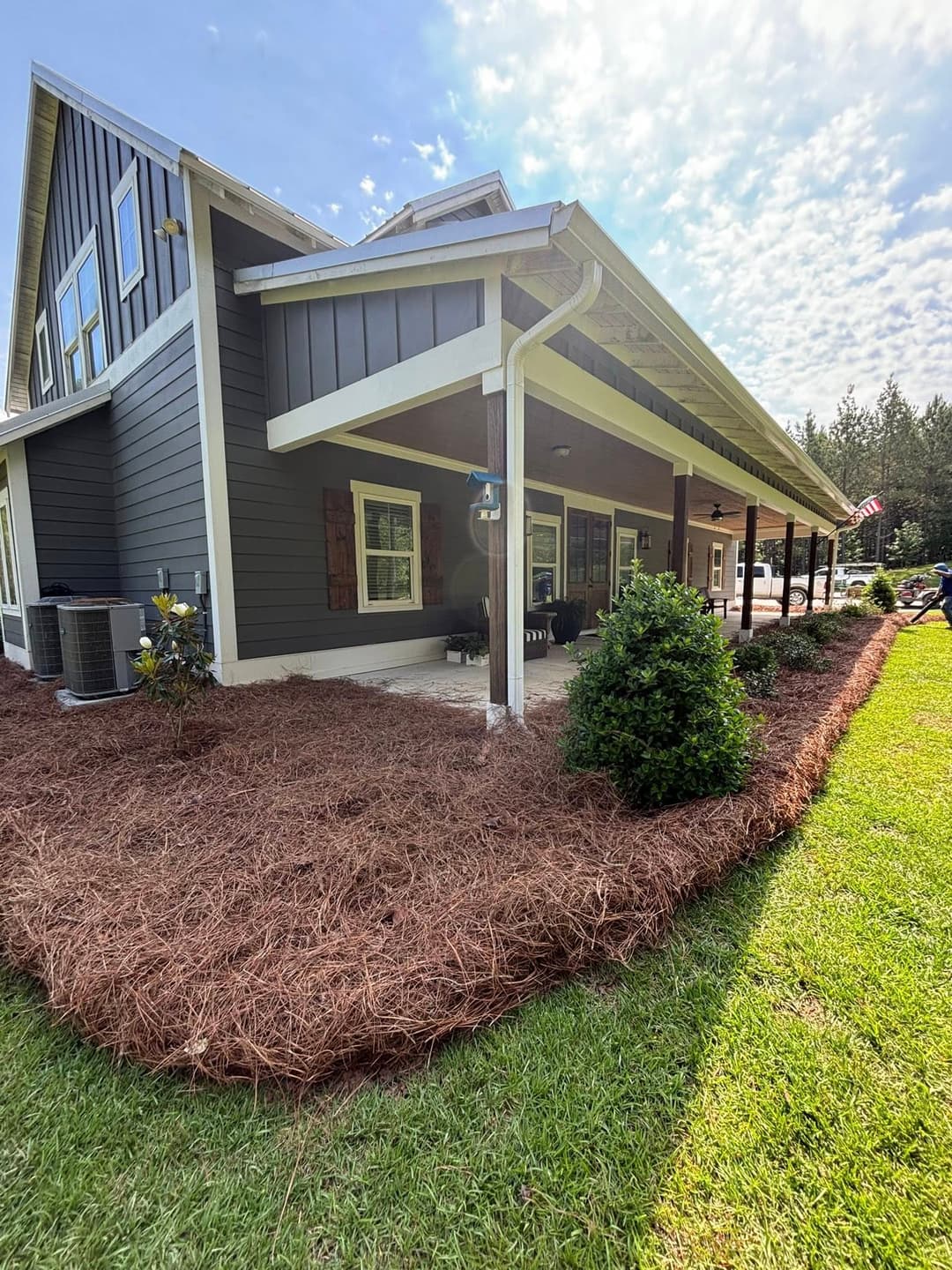 Modern house exterior with lush landscaping and pine straw mulch under a bright sky.