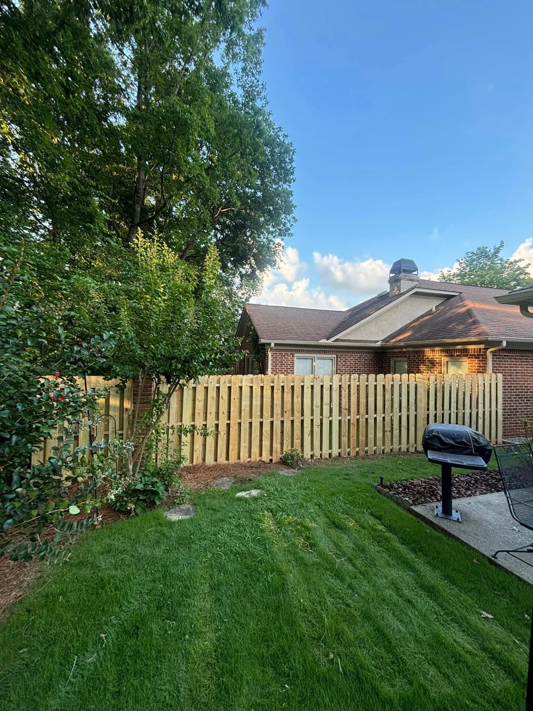 Lush backyard with green grass, wooden fence, and a grill near a brick house.