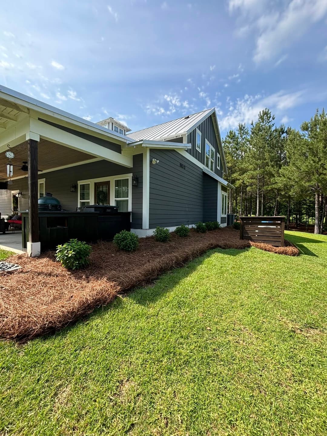 Side view of modern house with blue siding, landscaped yard, and pine trees in the background.