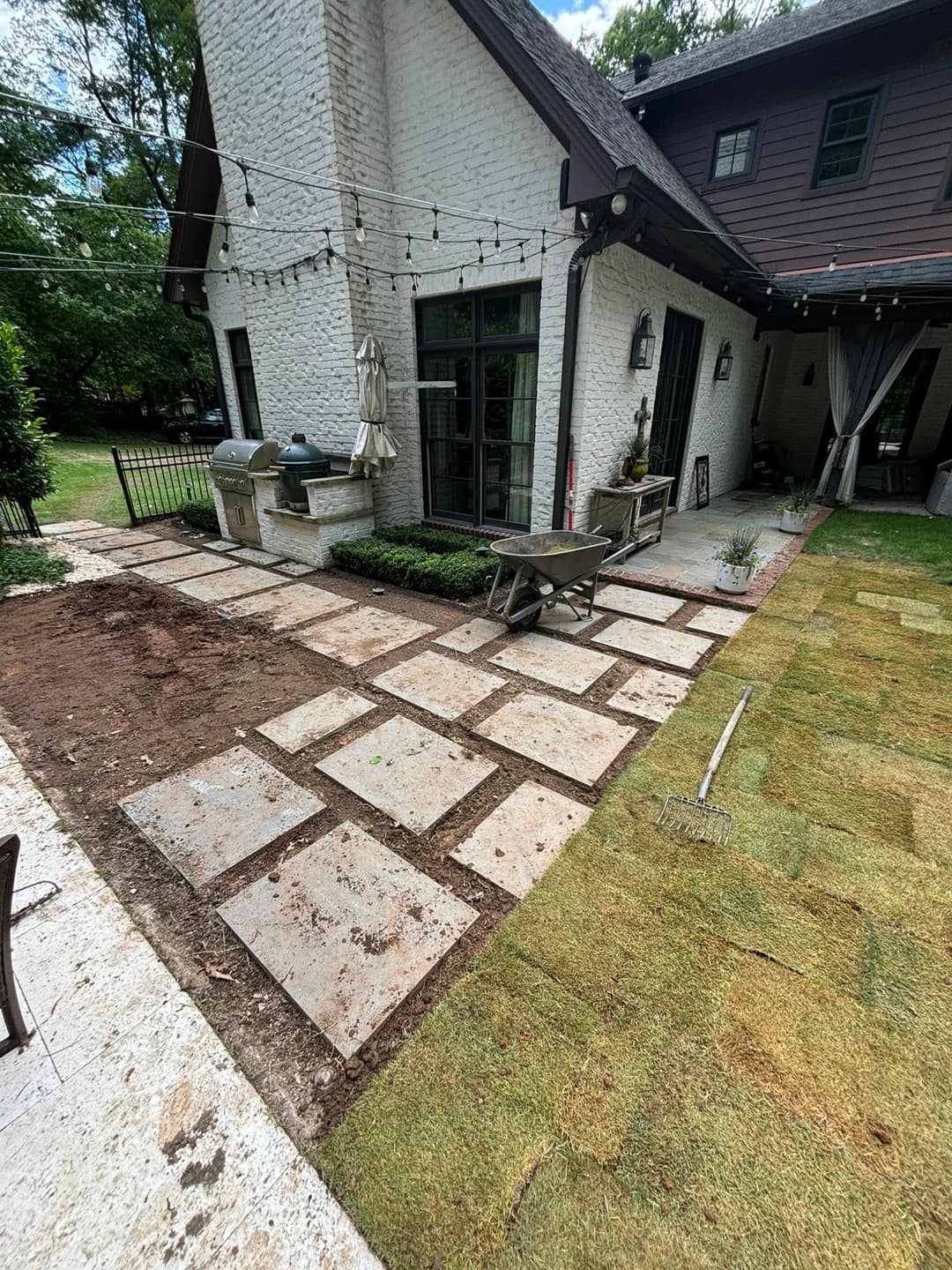 Backyard landscaping with stone pathway, wheelbarrow, and grill area in a residential setting.