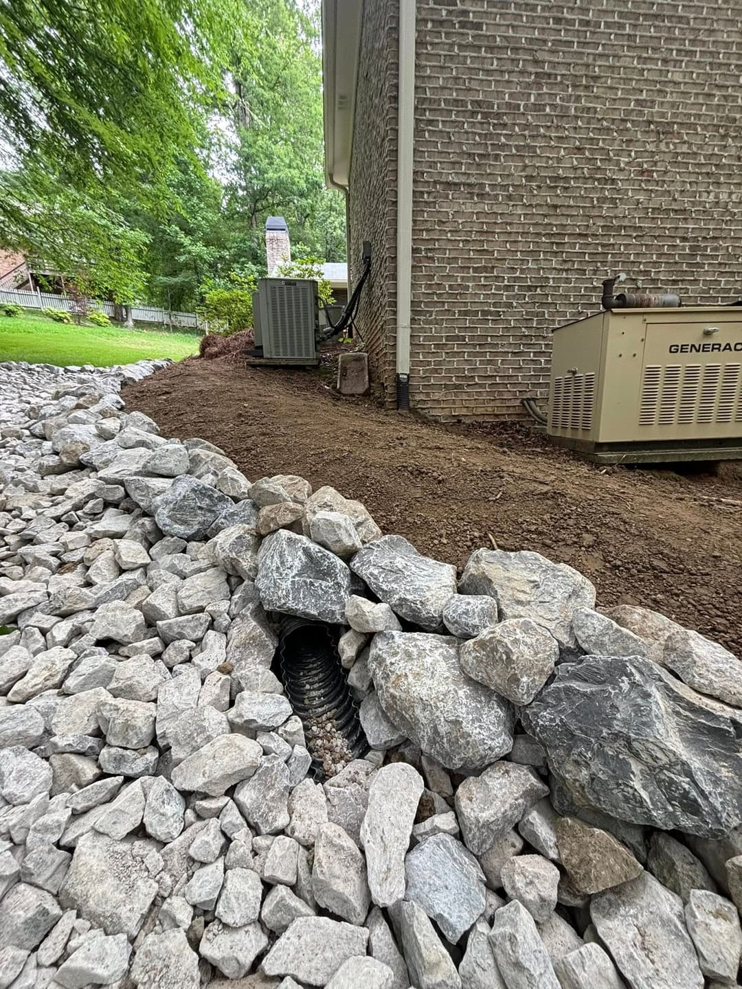 Drainage pipe surrounded by landscaping stones, beside a brick home and generator.