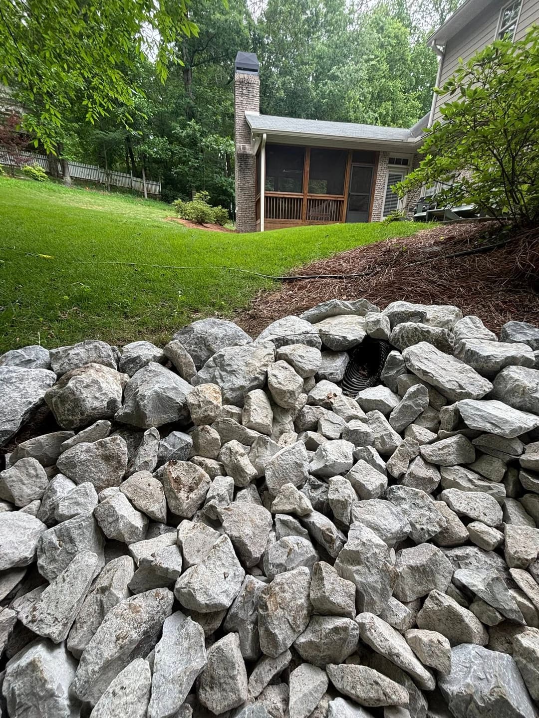 Landscape featuring a stone wall in the foreground with a house and greenery in the background.