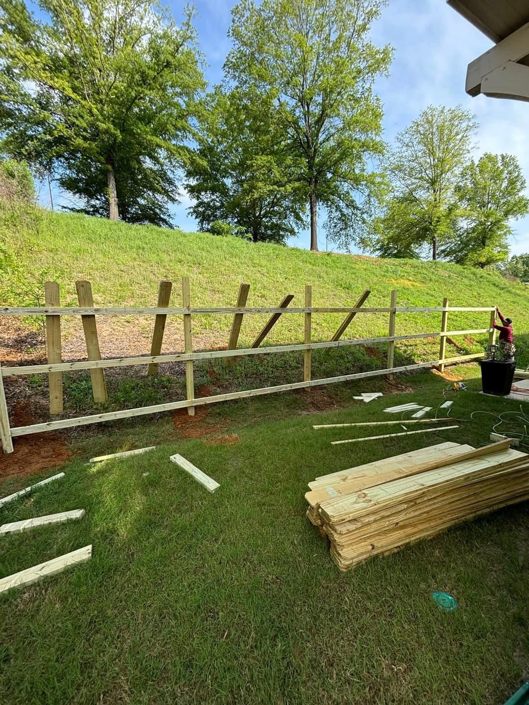 Wooden fence installation in a grassy area with trees and lumber scattered on the ground.