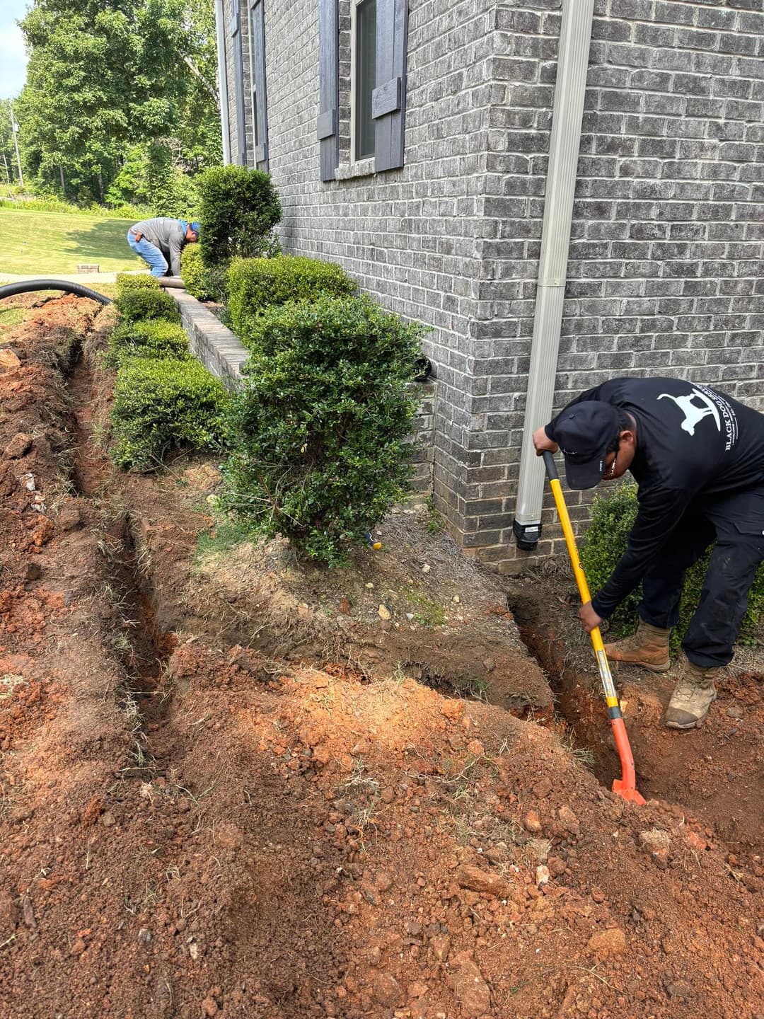 Workers digging a trench beside a brick house, preparing for drainage installation.