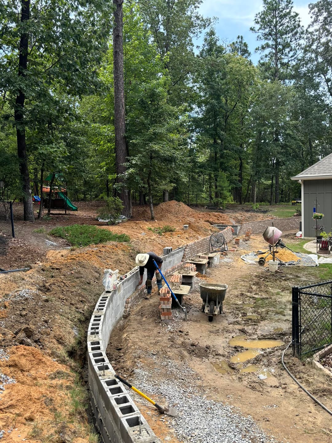 Worker laying a concrete block wall in a wooded backyard under construction.