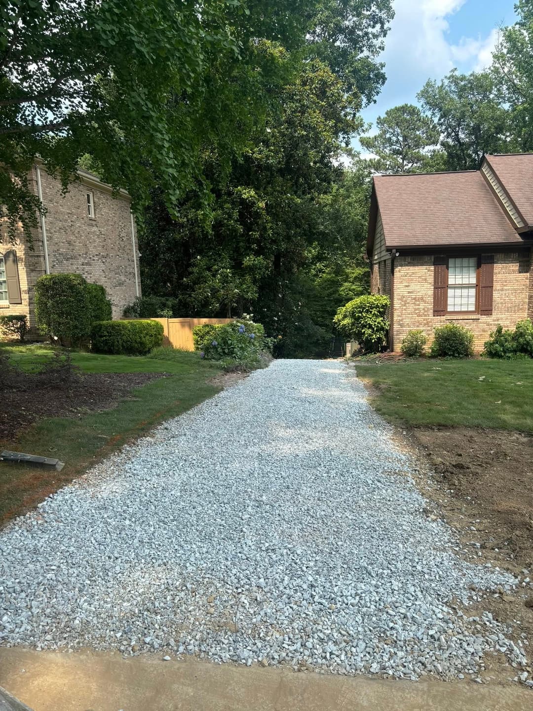 Gravel driveway installation between two homes with lush greenery and trees.
