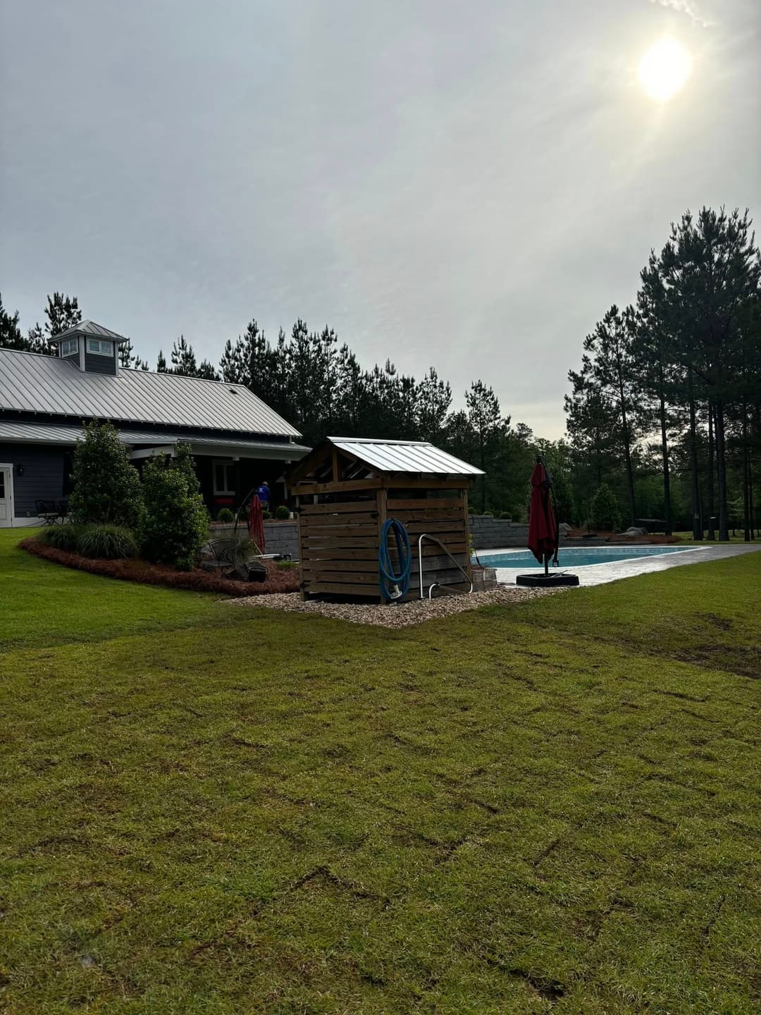 Wooden pool house with hoses near a swimming pool, surrounded by greenery and trees.