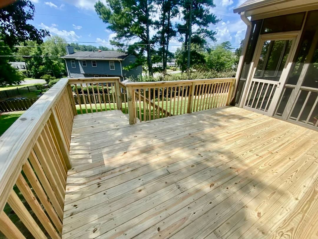 Spacious wooden deck with railing, surrounded by trees and residential view in daylight.