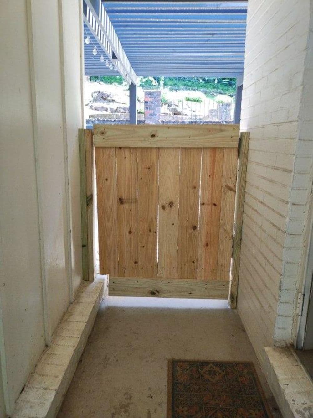 Wooden gate at the entrance of a home, framed by white walls and a decorative mat.