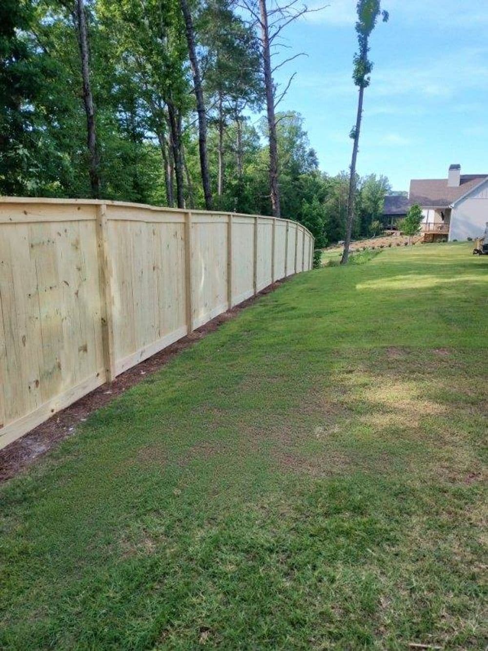 Wooden privacy fence curving along a green lawn bordered by trees and houses in the background.