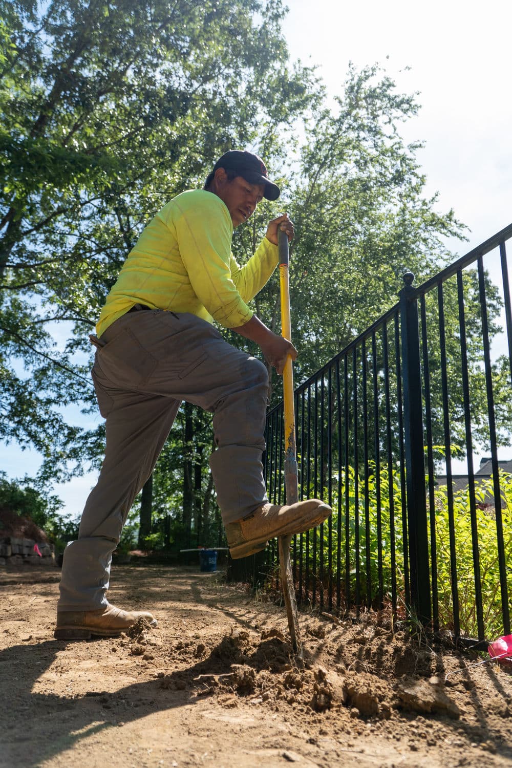 Worker digging soil next to a black iron fence in a landscaped area under a sunny sky.