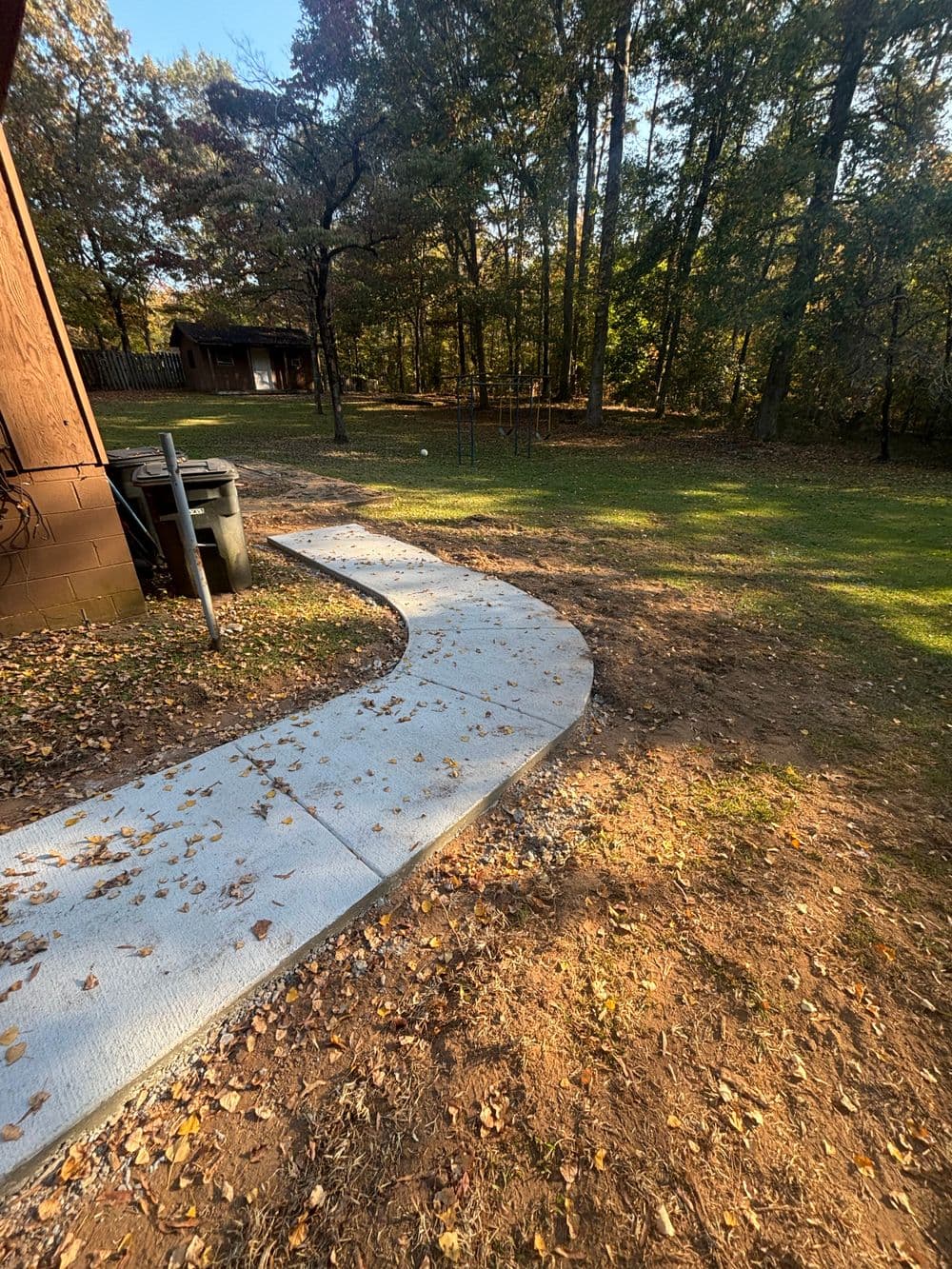 Curved concrete pathway leading through a wooded area with fallen leaves.