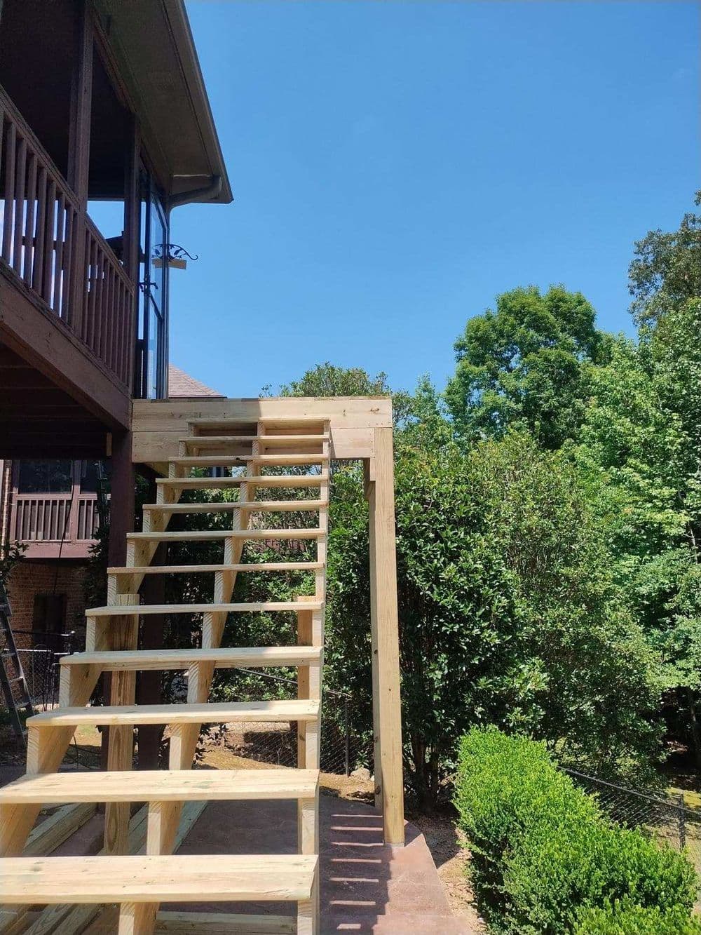 Wooden outdoor staircase leading to a house, surrounded by lush greenery and clear blue sky.
