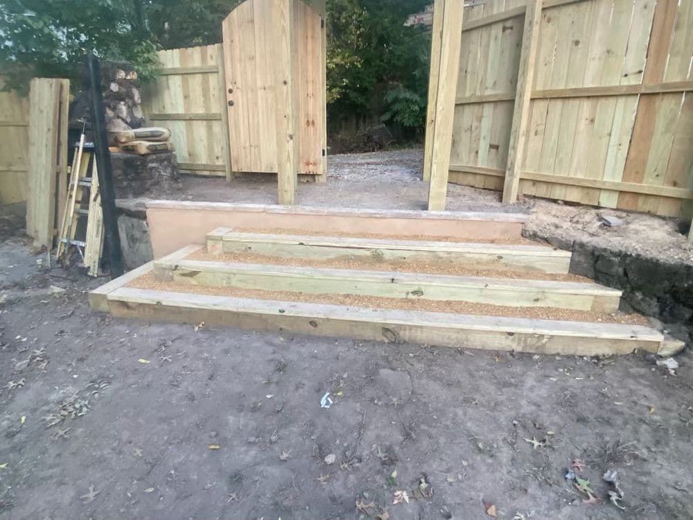 Wooden steps leading up to a gated entrance, framed by wooden fencing and gravel.