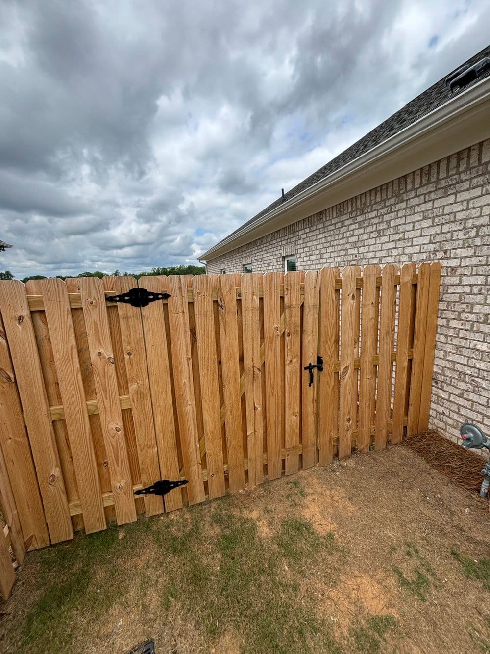 Wooden fence with black hardware, positioned next to a brick house under a cloudy sky.