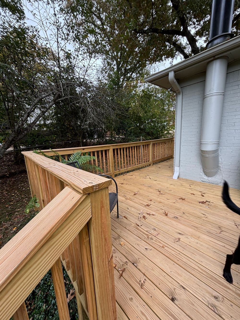 Wooden deck with railing, surrounded by trees and fallen leaves, near a chimney.