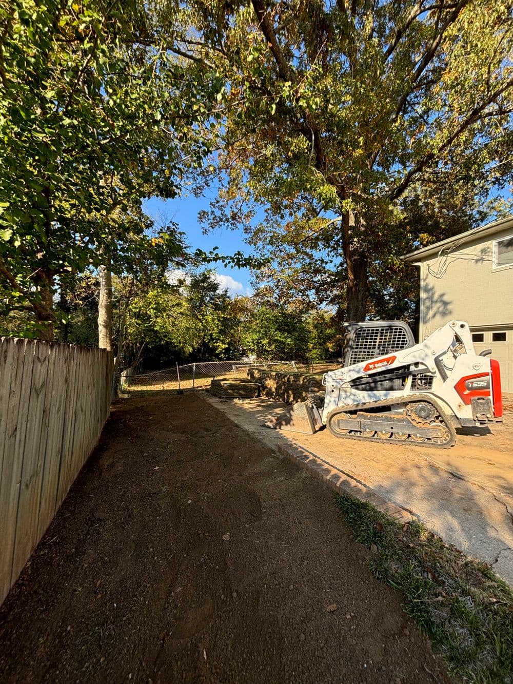 Bobcat parked on a dirt path in a wooded backyard, surrounded by trees and a wooden fence.