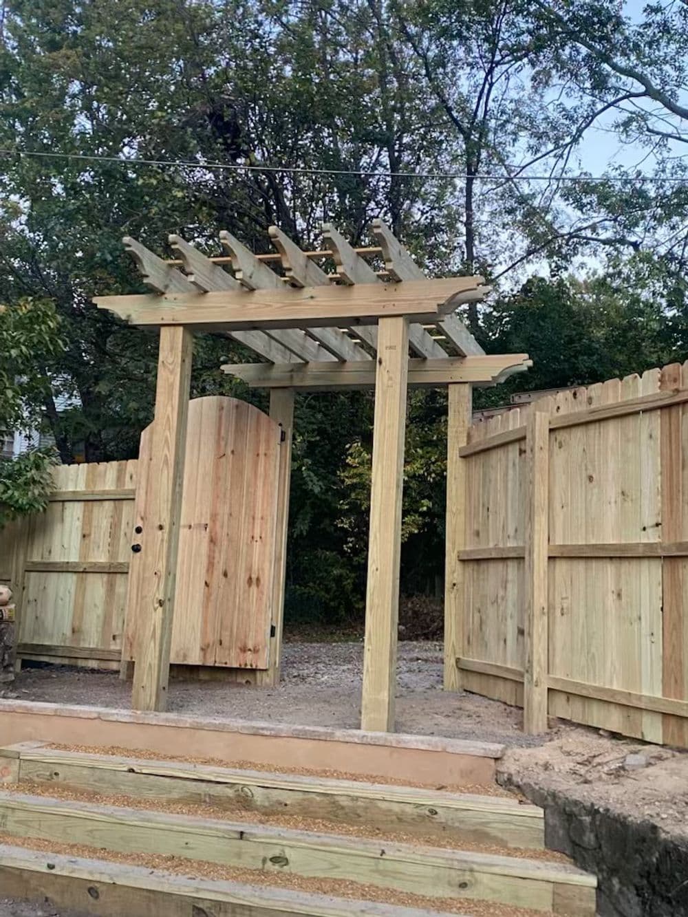 Wooden garden gate with pergola and surrounding fence in a landscaped yard.