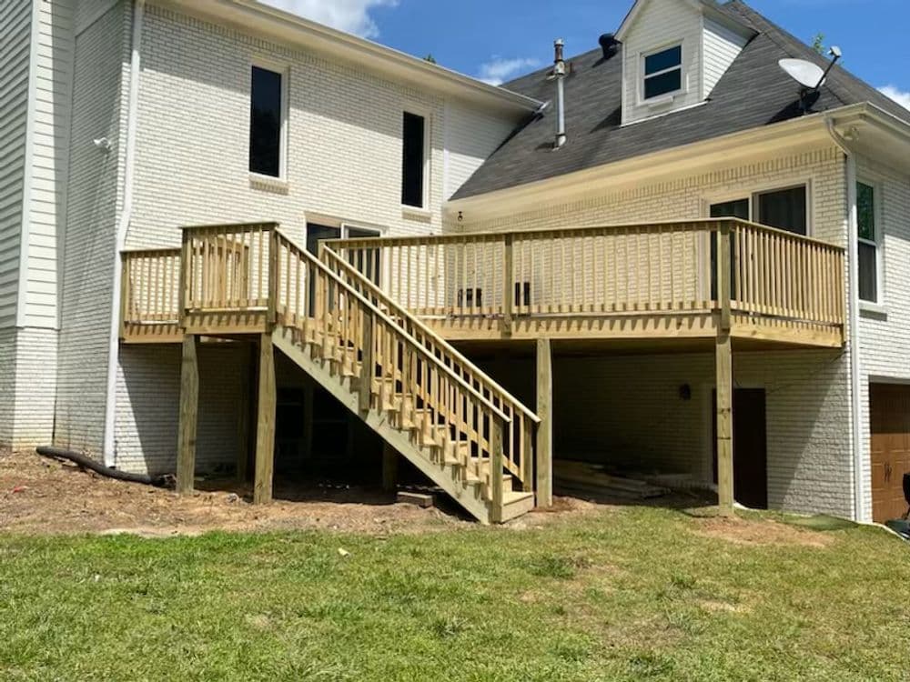 Wooden deck with stairs, extending from a white brick house, surrounded by grass.