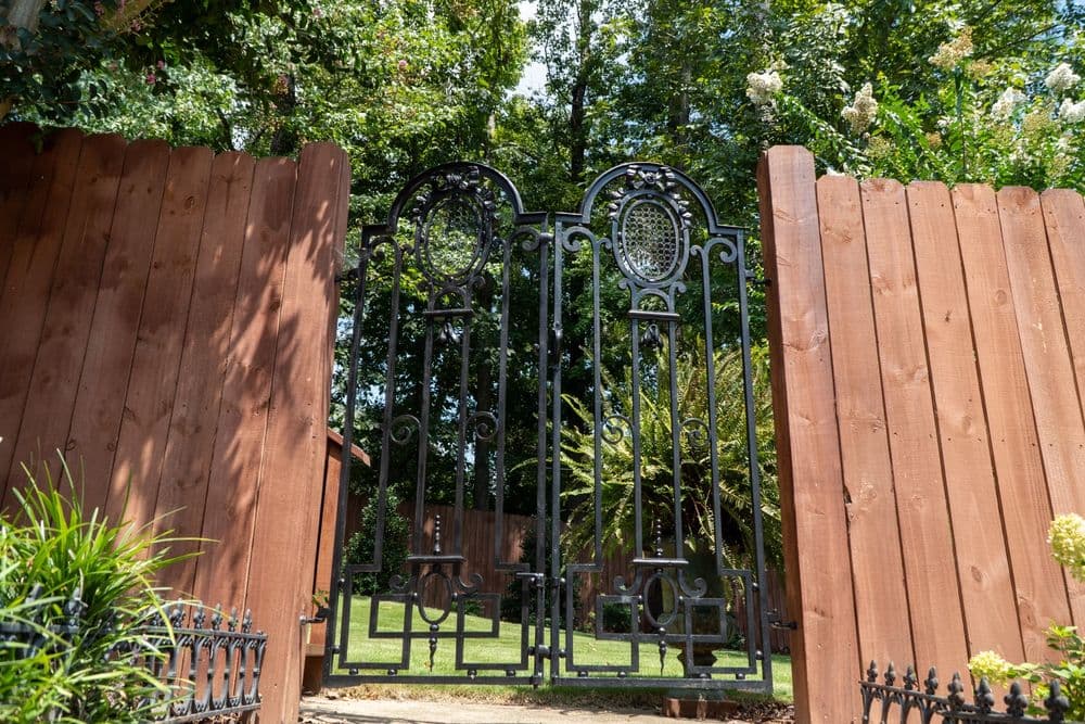 Ornate black iron gate opening to a lush green garden surrounded by wooden fencing.