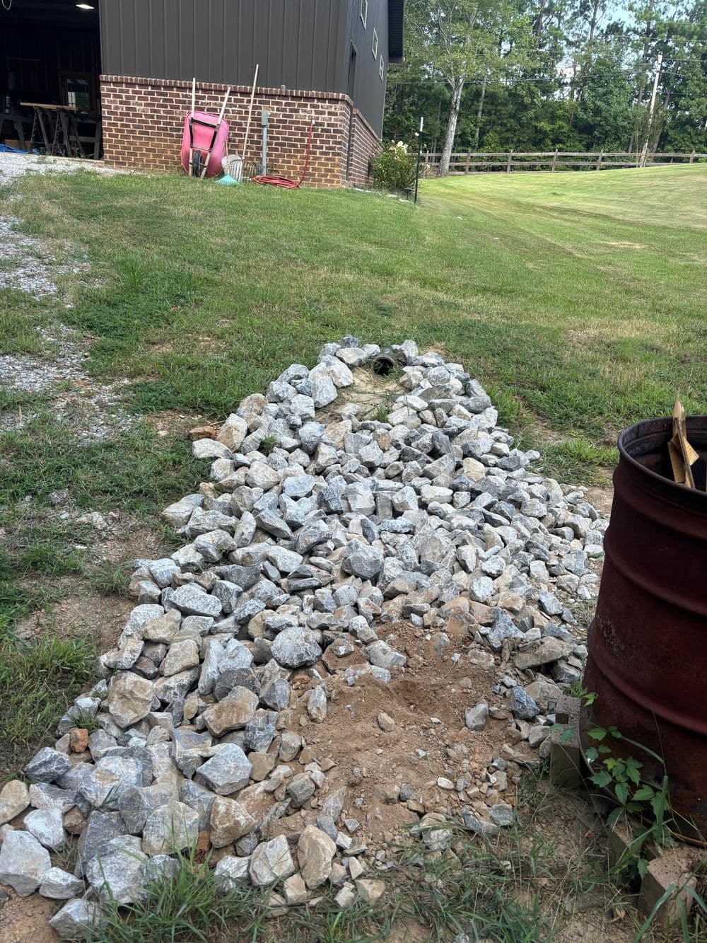 Piled rocks in a yard near a barn, with grass and a metal barrel nearby.