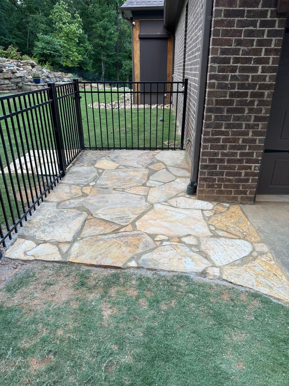 Flagstone patio area next to a house, enclosed by a black metal fence. Green lawn in background.