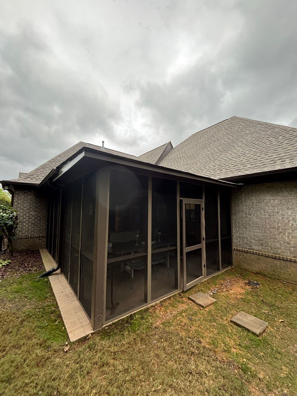Screened-in porch attached to a brick house, surrounded by grass and cloudy sky.