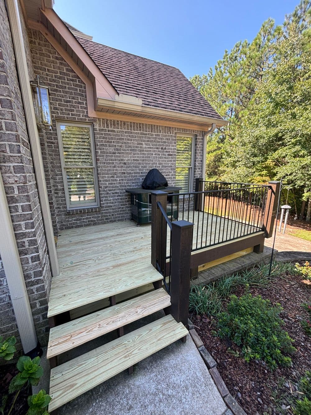 Backyard deck with steps, grill covered, surrounded by greenery and trees on a sunny day.