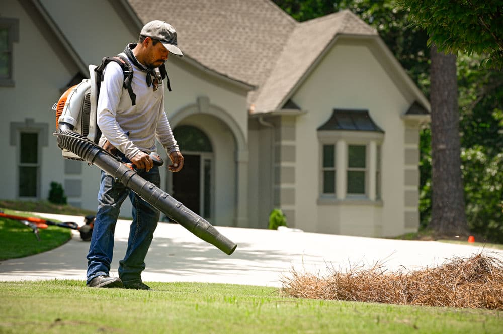 Man using a backpack leaf blower to clear debris from a landscaped yard near a home.